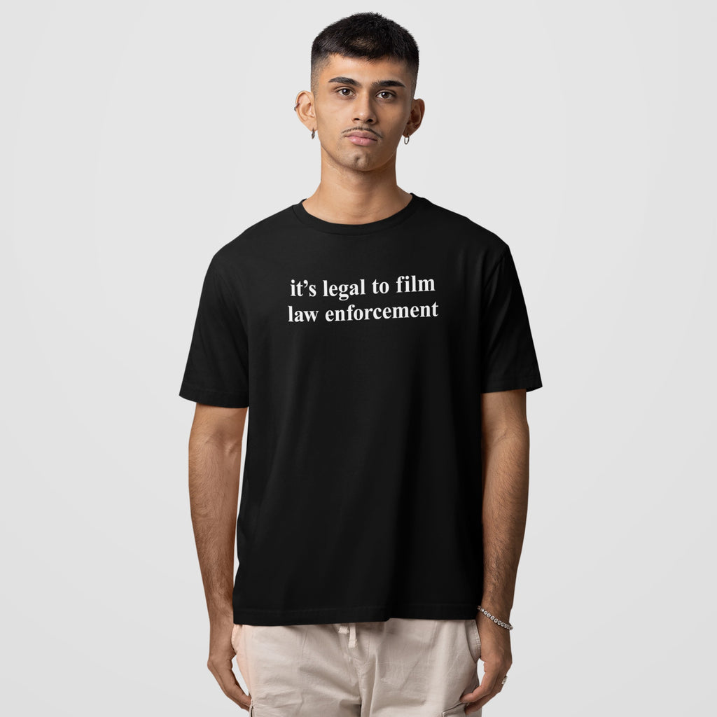 Young man wearing a black Film The Police T-shirt with it's legal to film law enforcement text, paired with light-colored casual pants, embodies a message of activism and awareness.