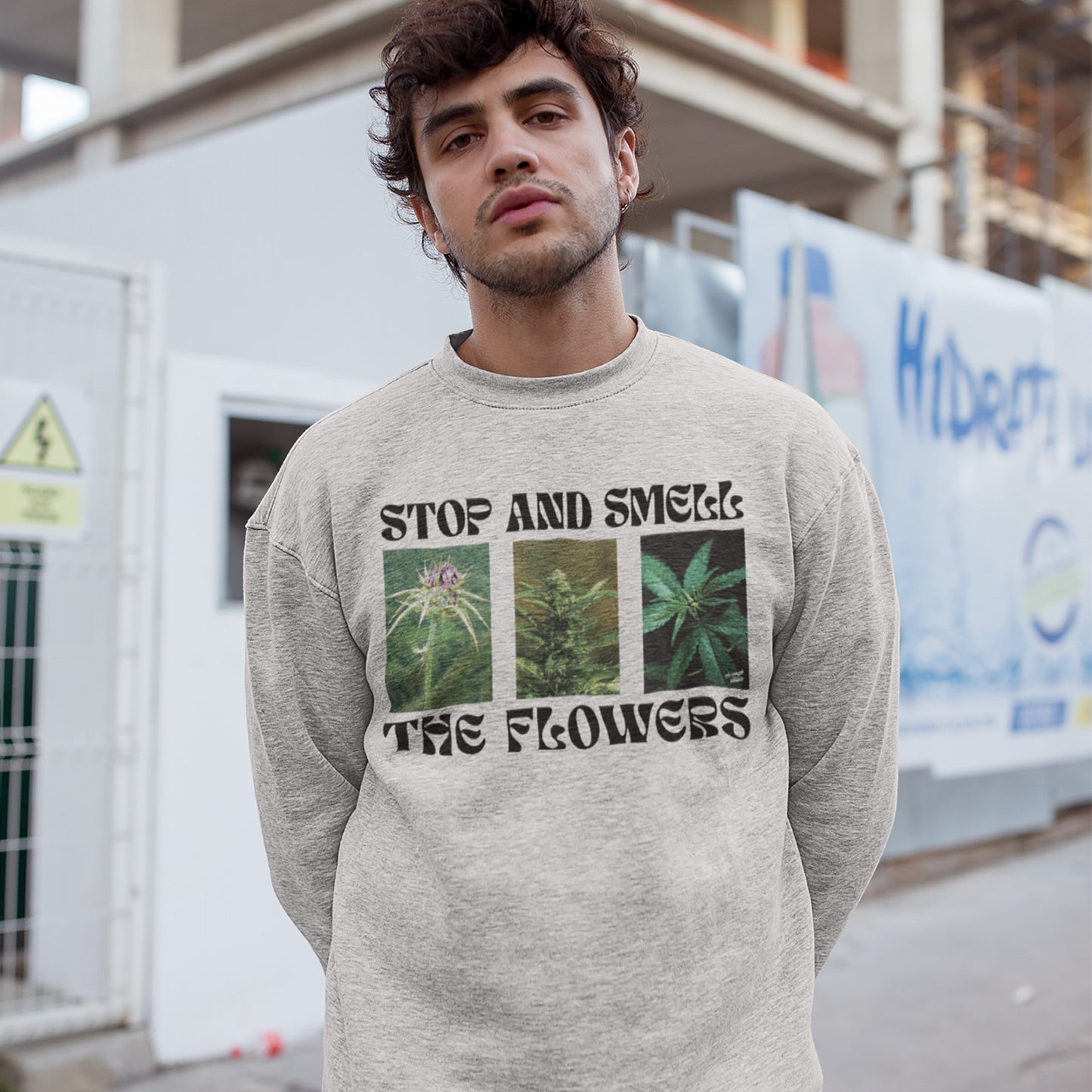 Young man wearing the Flowers Hoodie & Sweatshirt with cannabis plant images and Stop and Smell the Flowers text, standing in front of a construction site.