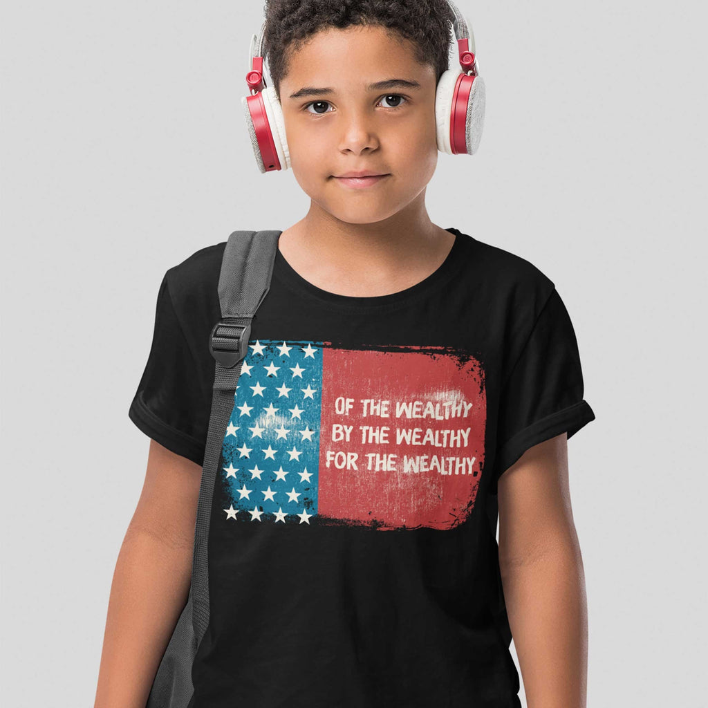 A pre-teen wears the For The Wealthy T-shirt featuring a distressed American flag and protest slogan, paired with white and red headphones and a backpack strap over one shoulder.