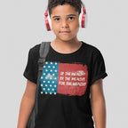 A pre-teen wears the For The Wealthy T-shirt featuring a distressed American flag and protest slogan, paired with white and red headphones and a backpack strap over one shoulder.