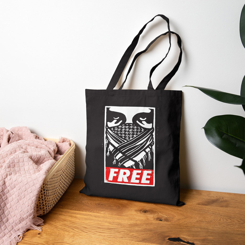 Free Palestine JULZ Tote Bag featuring a masked figure graphic and bold FREE text. Positioned on a tabletop beside a pink throw in a basket and a potted plant.