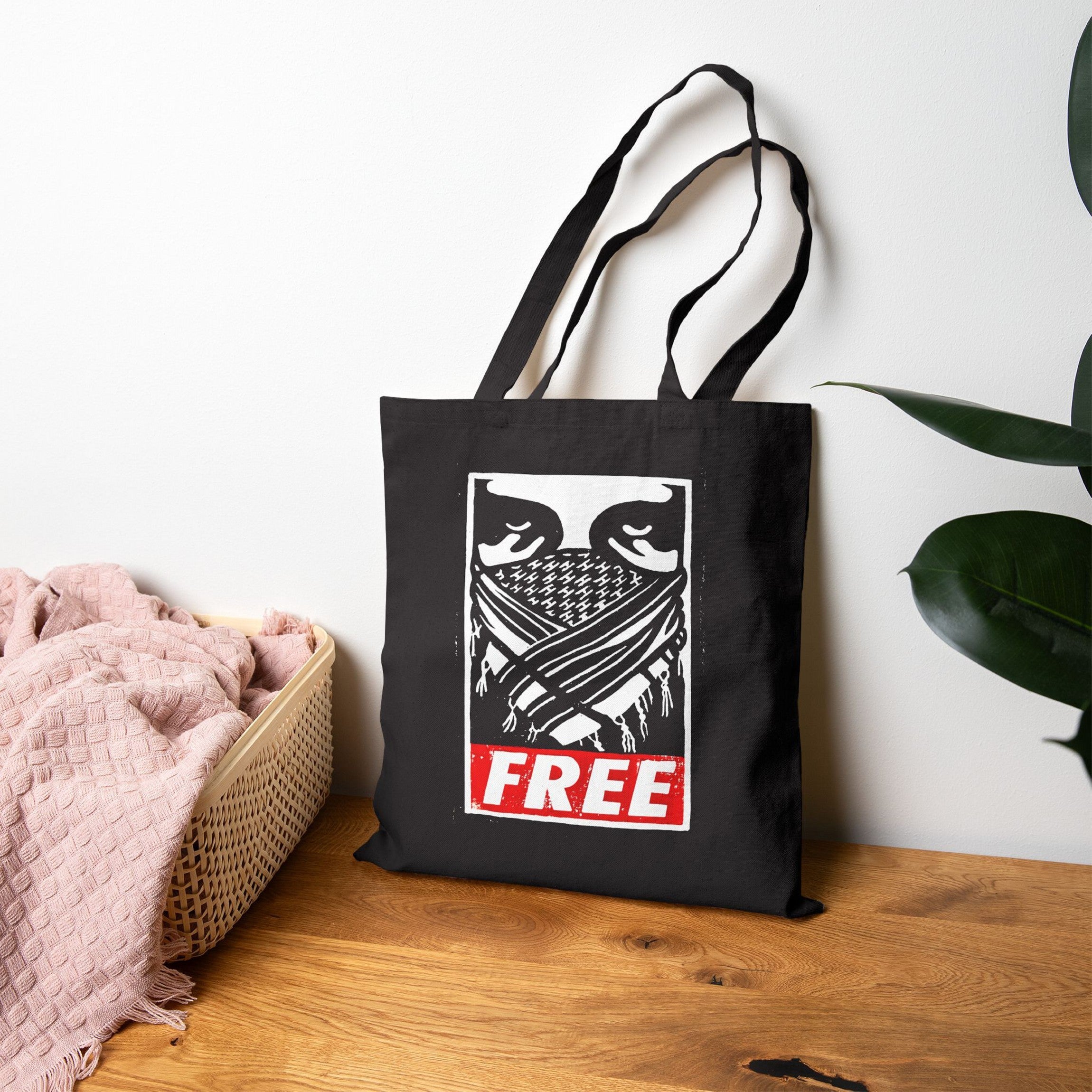 Free Palestine JULZ Tote Bag featuring a masked figure graphic and bold FREE text. Positioned on a tabletop beside a pink throw in a basket and a potted plant.