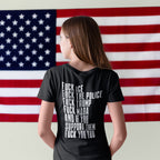 Person facing a large American flag wearing the Fuck 'Em All Back Print Shirt with bold protest text against ICE, police, Trump, and MAGA. Unisex fit, ethically made for resistance.