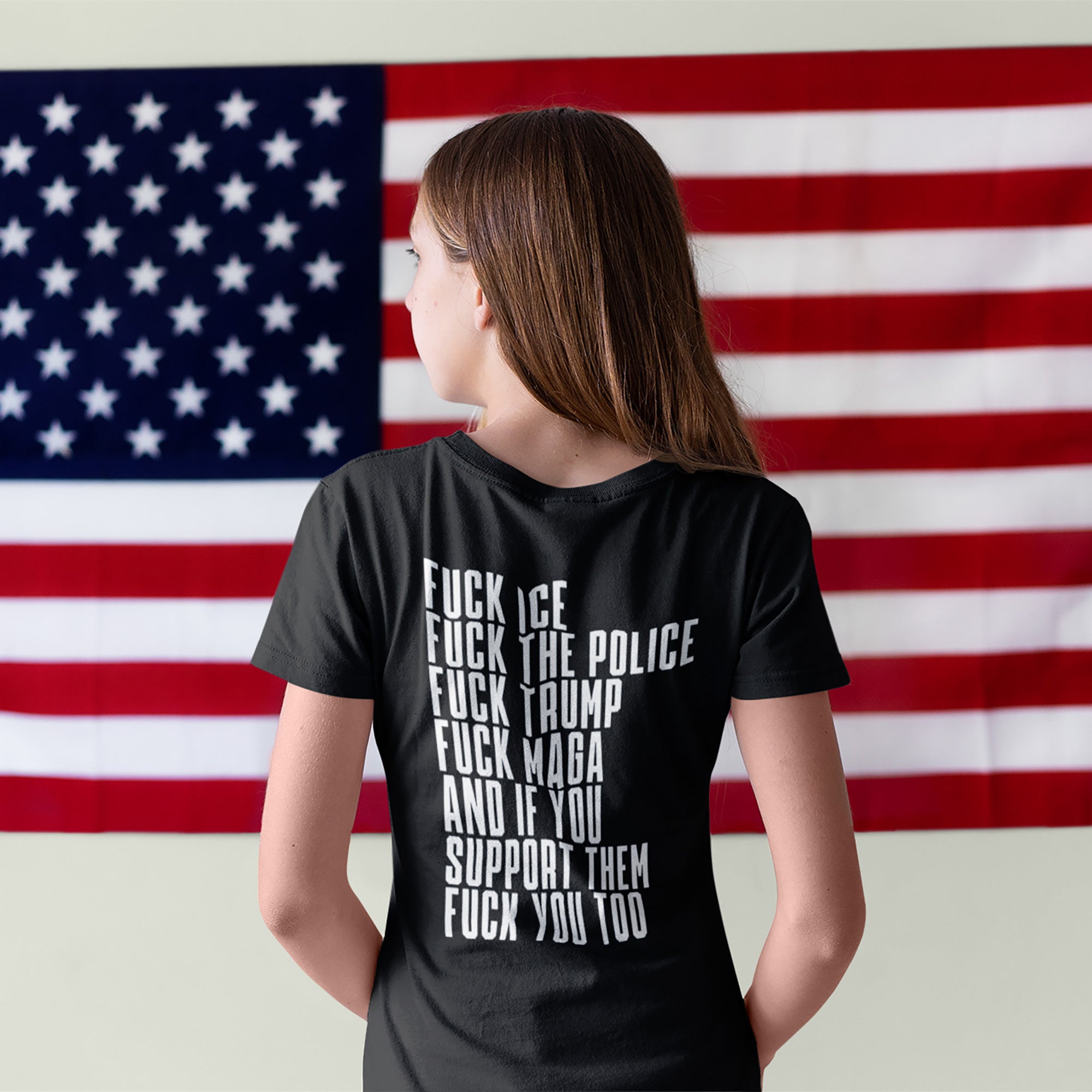 Person facing a large American flag wearing the Fuck 'Em All Back Print Shirt with bold protest text against ICE, police, Trump, and MAGA. Unisex fit, ethically made for resistance.