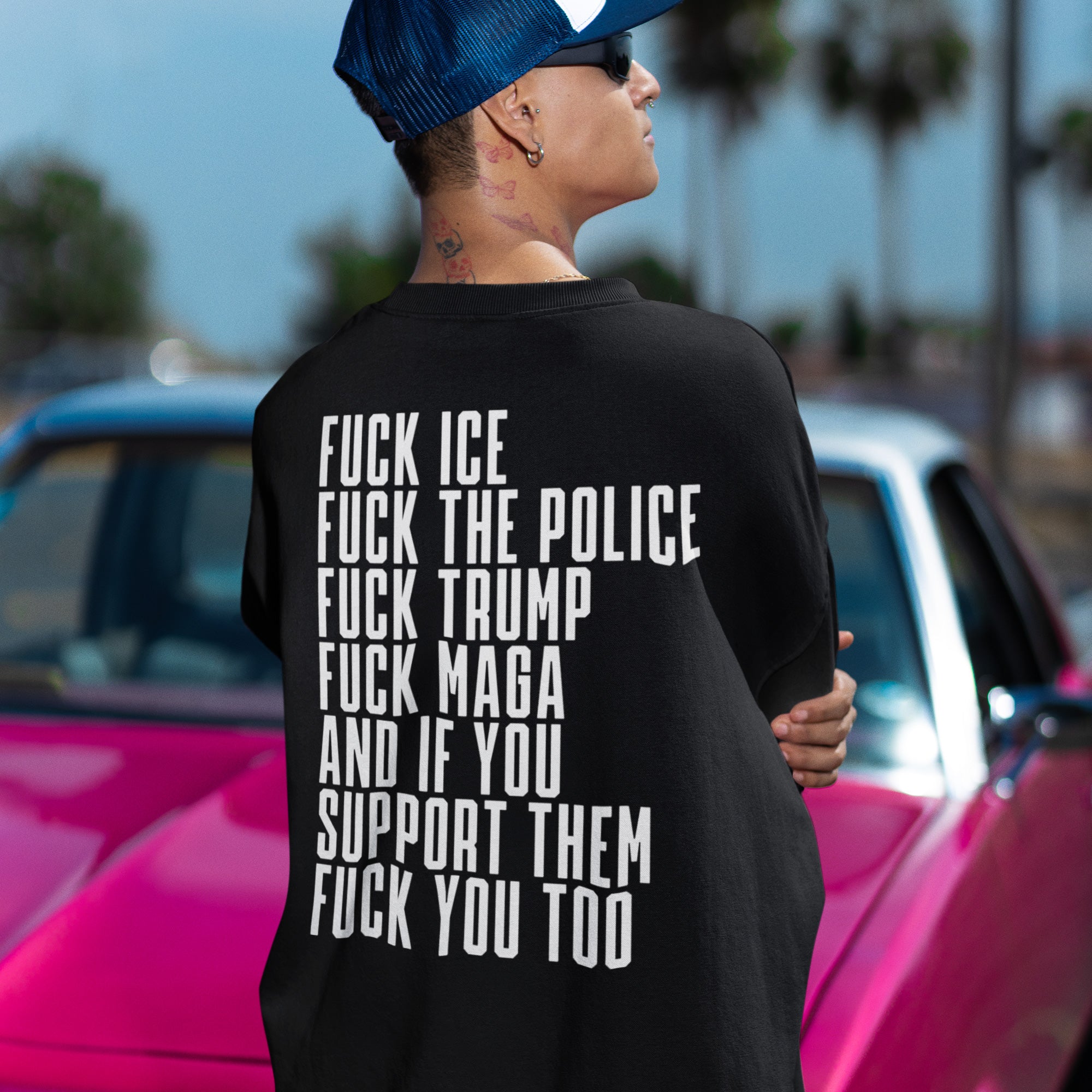 Person wearing a Fuck 'Em All Back Print Shirt featuring bold protest messages, standing in front of a pink car, embodying anti-fascist and resistance themes with impactful statements.