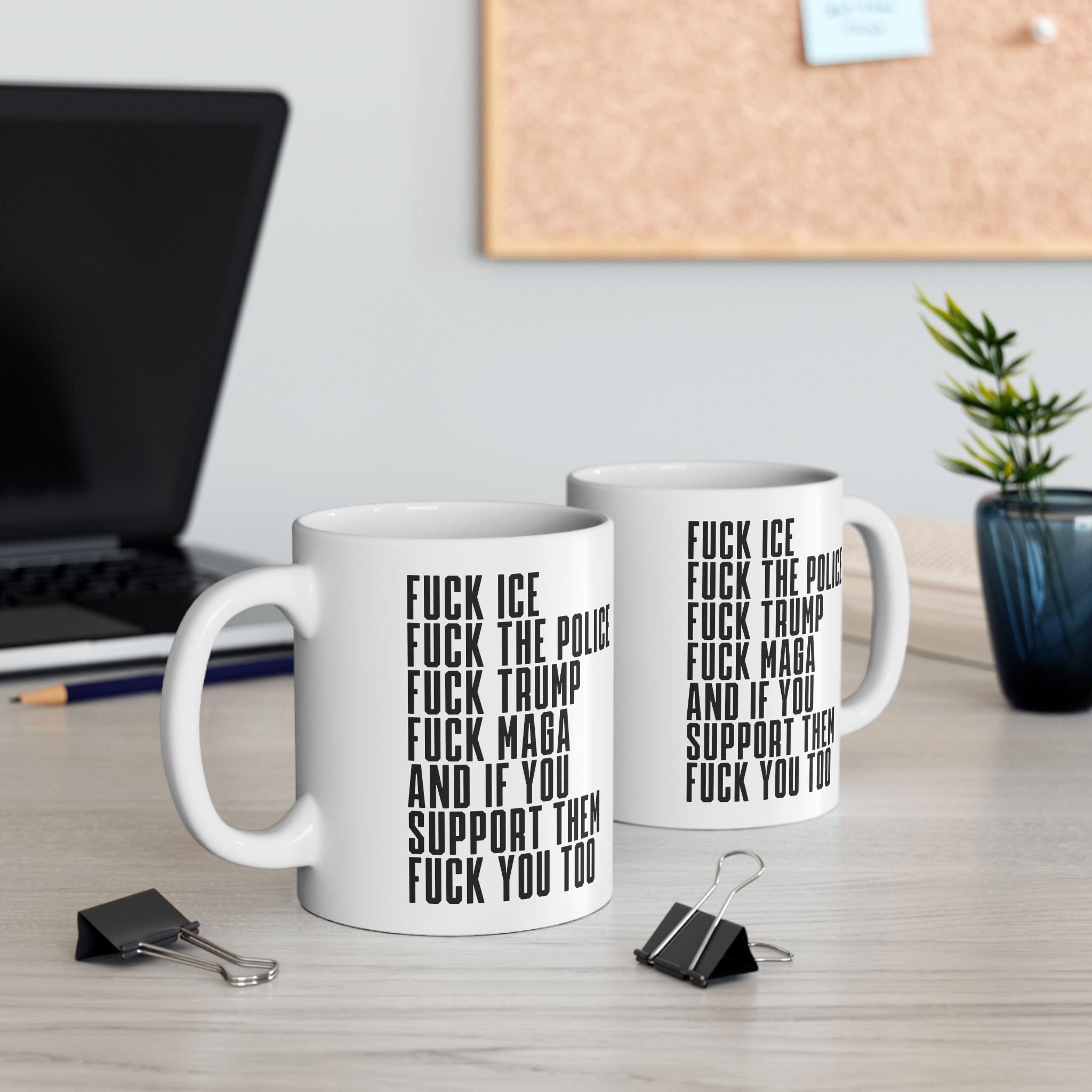 The Fuck 'Em All Mug features bold anti-fascist slogans on white ceramic, ideal for making a statement. Shown on a desk with office items and a potted plant nearby.