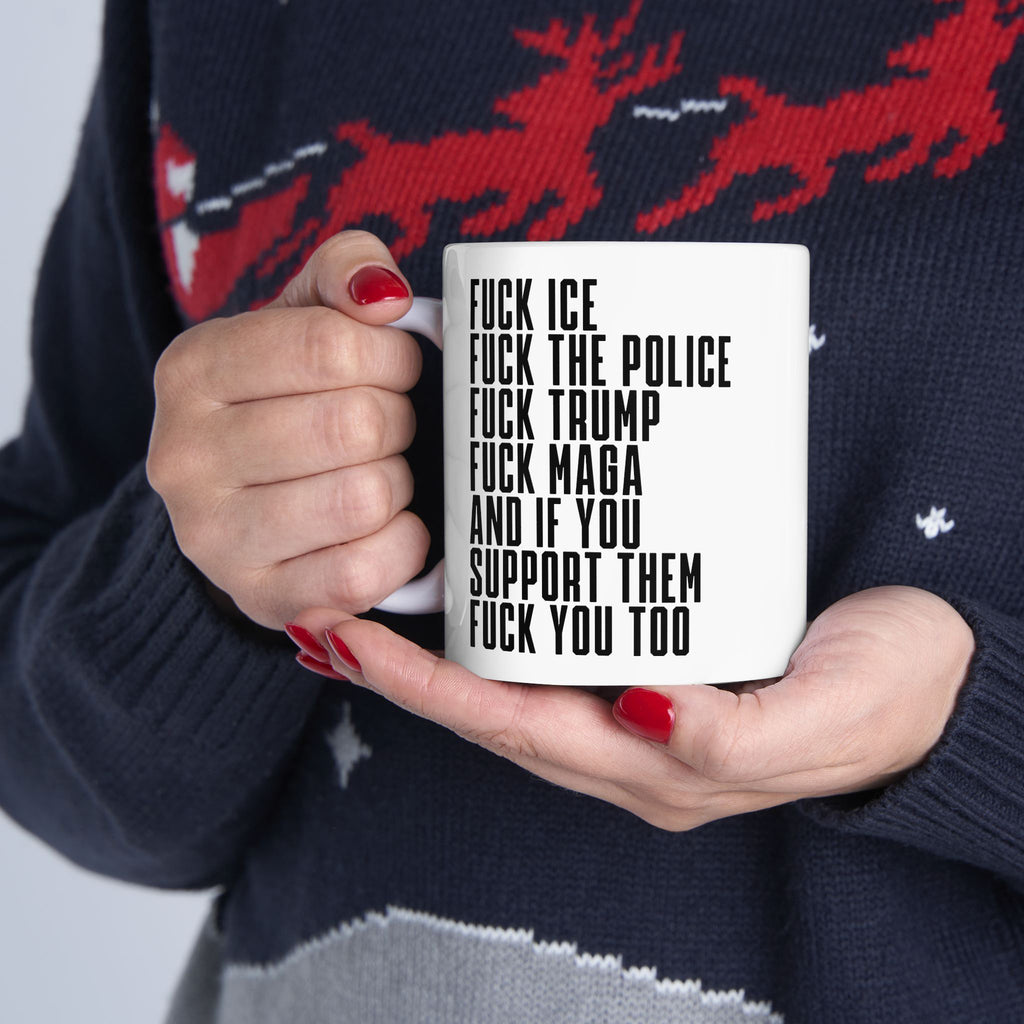 Person holding the Fuck 'Em All Mug, featuring bold, profanity-laden political slogans in black on a white ceramic surface. The holder has red nails and wears a dark sweater with a reindeer motif.