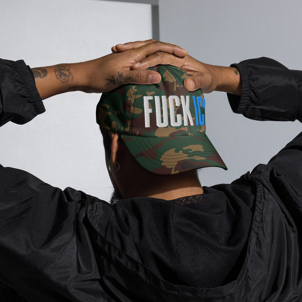 Back view of a person wearing a woodland-camouflage Fuck ICE Hat, featuring bold 3D puff embroidery, embodying defiance and visibility, with tattooed forearms and dressed in a black windbreaker.