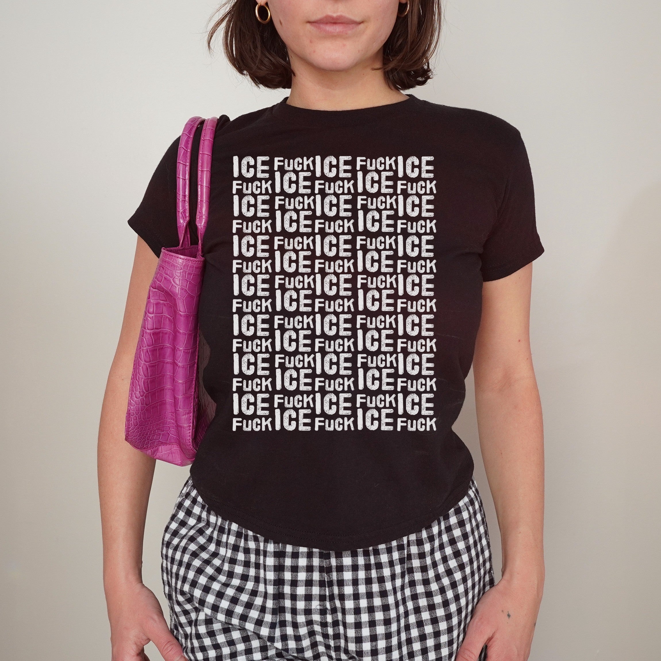 Person wearing the Fuck ICE Pattern Women's Baby Tee featuring a bold white-text grid design, paired with black-and-white gingham pants and a purple shoulder bag, cropped from shoulders to mid-thigh.