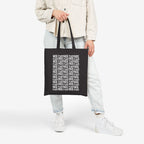 Person holding a black Fuck ICE Pattern Tote Bag, featuring bold white text pattern. Paired with casual light-wash jeans, cream shacket, and white sneakers, symbolizing protest and advocacy through everyday style.