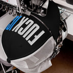Trucker-style Fuck ICE hat in embroidery machine, stitching visible, with white mesh back and FUSION in progress, showcasing bold values and expressive design.