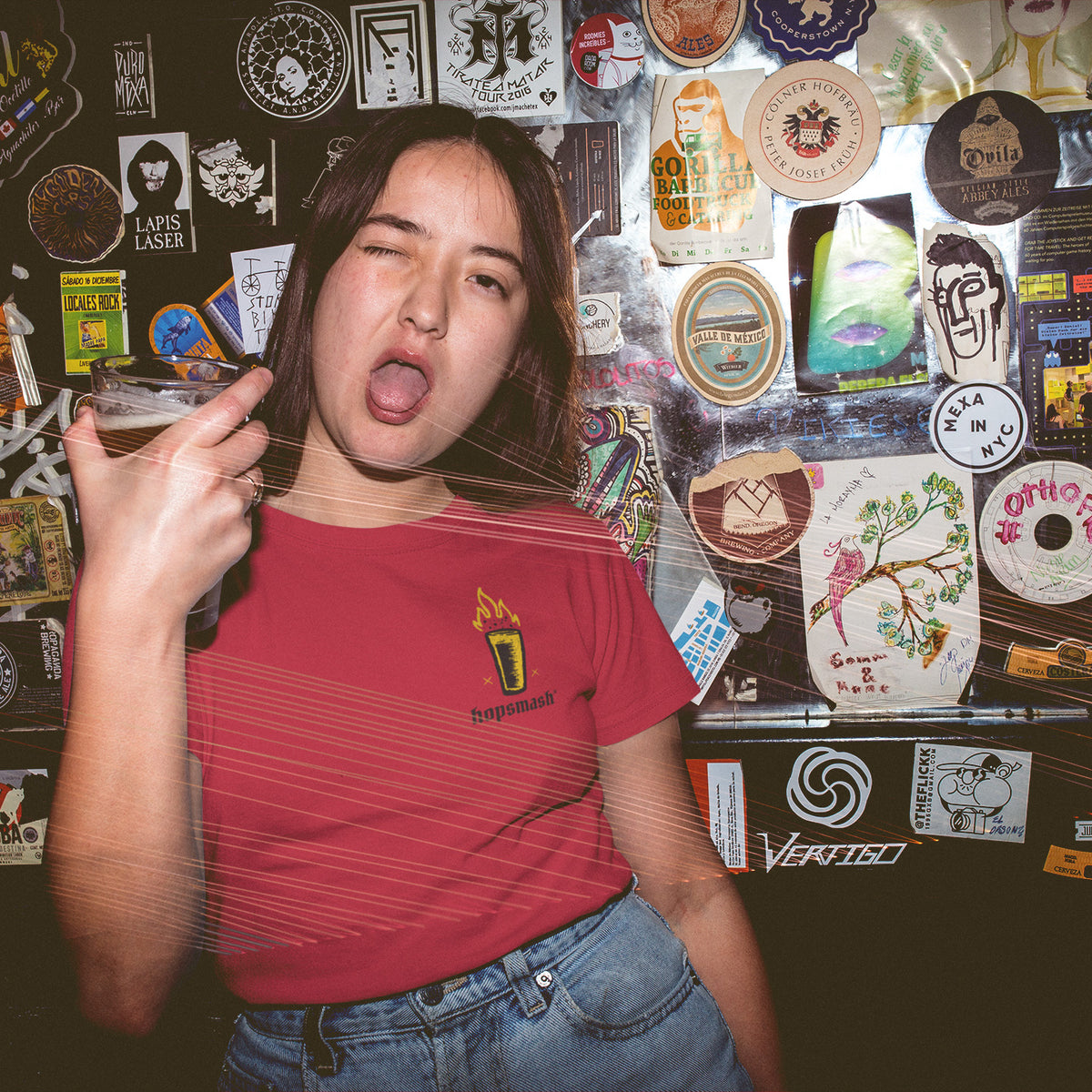 A woman wearing a red t-shirt with a small yellow and black graphic Hopsmash logo on the left chest, posing in front of a bar wall covered in stickers.