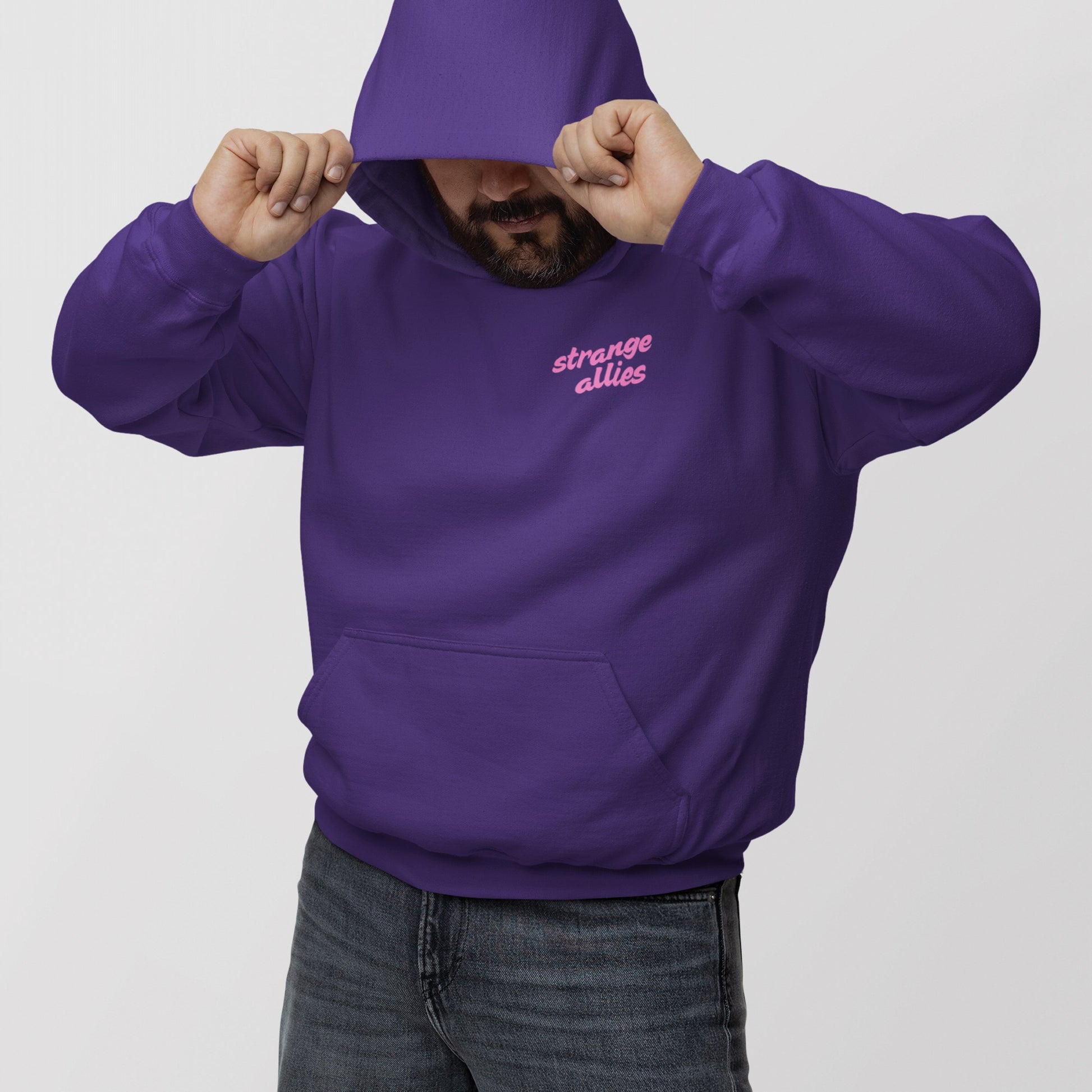 Person wears deep purple God Loves Ugly Hoodie & Sweatshirt, featuring strange allies in pink script. They pull the hood down slightly, partially obscuring their face, paired with dark-wash jeans.