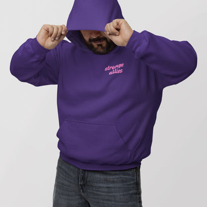 Person wears deep purple God Loves Ugly Hoodie & Sweatshirt, featuring strange allies in pink script. They pull the hood down slightly, partially obscuring their face, paired with dark-wash jeans.