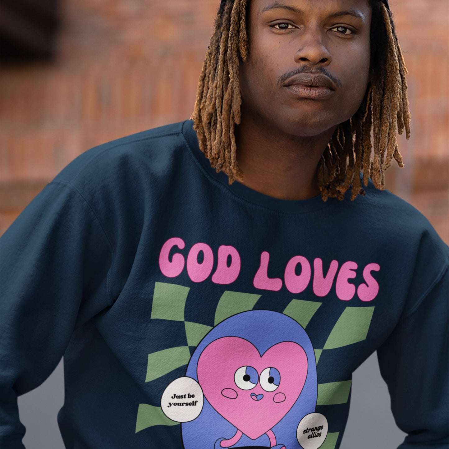 Young man with shoulder-length dreadlocks wears the God Loves Ugly Hoodie & Sweatshirt, featuring a colorful graphic with a heart character and speech bubbles, promoting individuality and defying perfection culture.