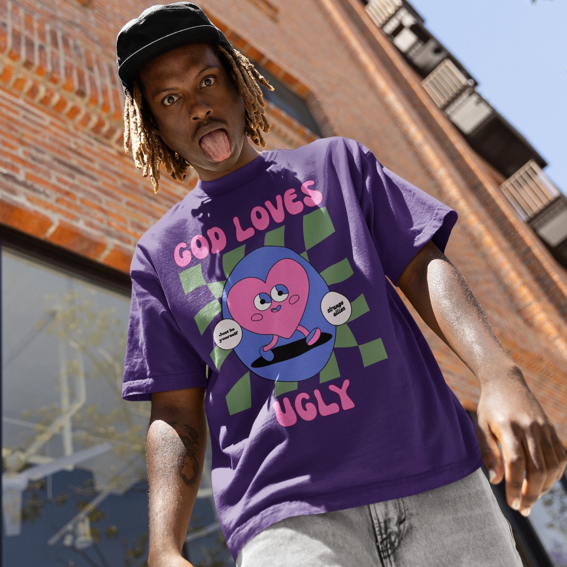 God Loves Ugly T-shirt featuring a cartoon pink heart skateboarding with affirmations “Just be yourself” and “strange allies.” The young man models it, embodying bold self-expression and inclusivity.