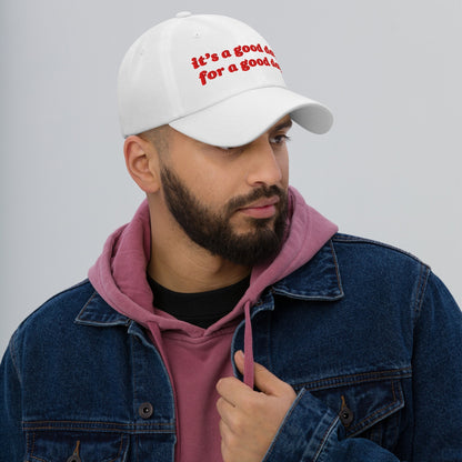 Young man wearing the Good Day Hat in white with red embroidery, paired with a dusty-rose hoodie and denim jacket. The hat promotes optimism and supports Amnesty International through purchases.