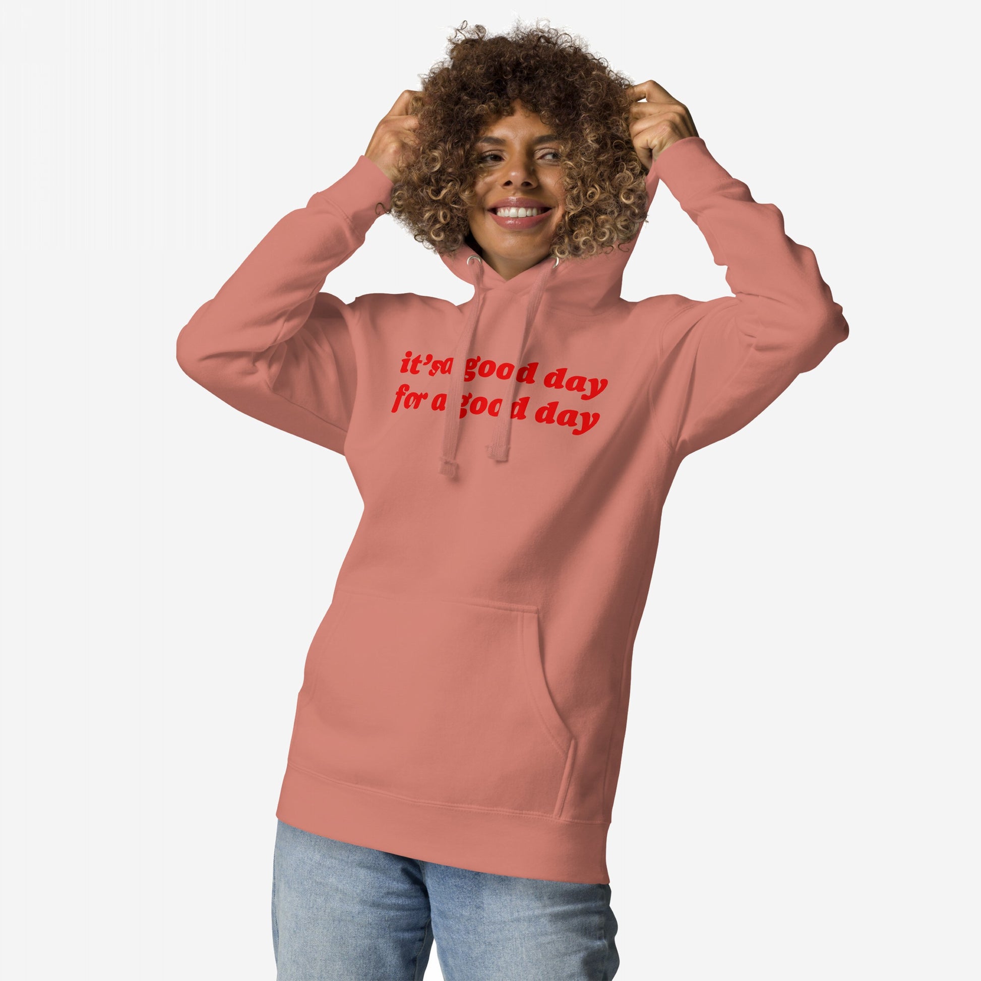 Smiling person wearing the Good Day Hoodie & Sweatshirt, showcasing its bold it’s a good day for a good day message, pulling the hood playfully, paired with light-wash jeans against a plain background.