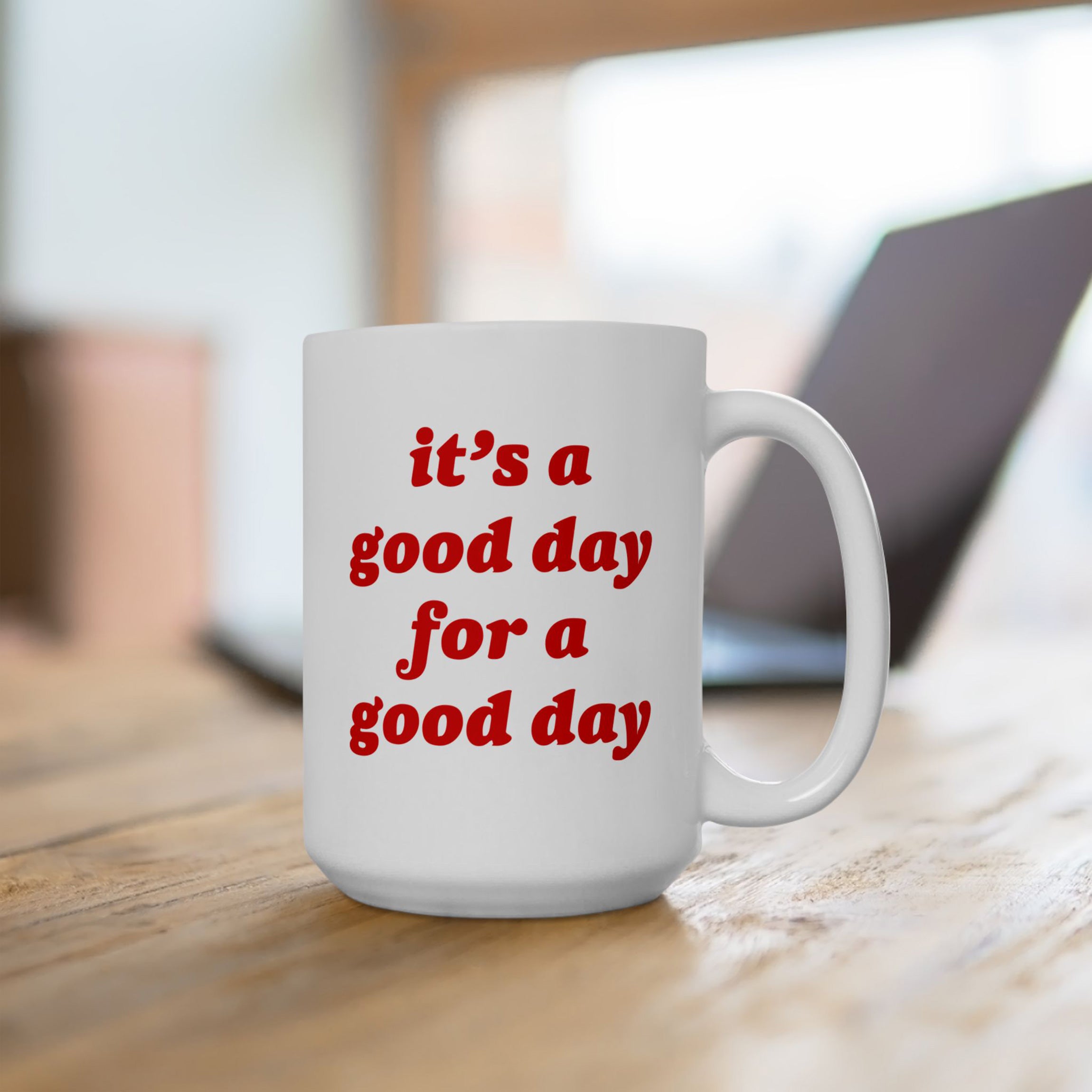 White ceramic Good Day Mug on wooden tabletop, featuring bold red text: it’s a good day for a good day. Positioned in front of a softly blurred laptop, emphasizing its playful and motivational vibe.