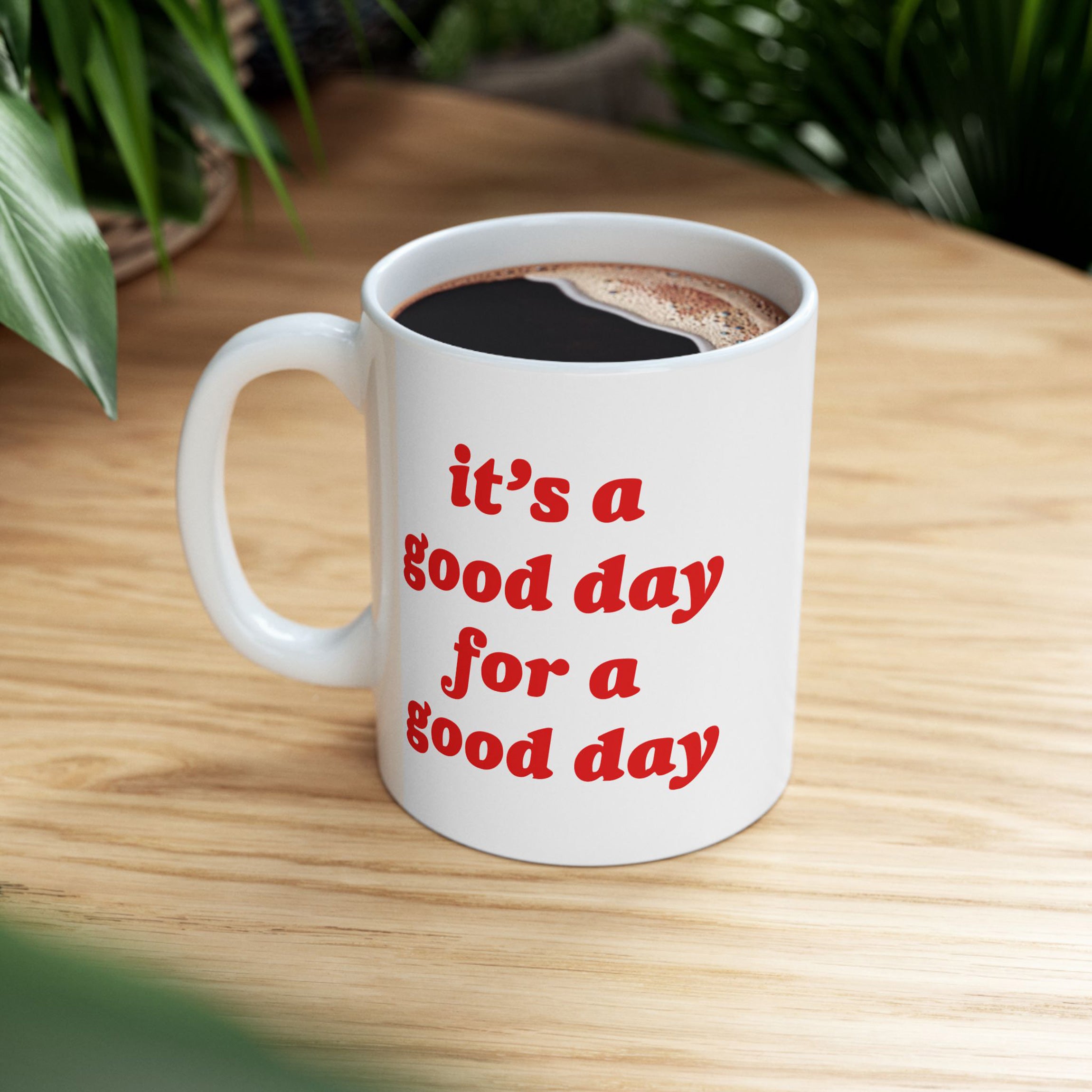 White ceramic Good Day Mug filled with black coffee, featuring bold red text it’s a good day for a good day, sits on a wooden tabletop, surrounded by green houseplants.