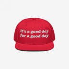 The Good Day Hat features white embroidered text stating it’s a good day for a good day on a red, flat-brim cap, promoting positivity and mental health advocacy with stylish, uplifting flair.