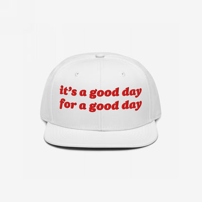 The Good Day Hat features a white, flat-brimmed baseball cap with red, retro-style embroidery stating “it’s a good day for a good day,” embodying optimism and support for mental health advocacy.