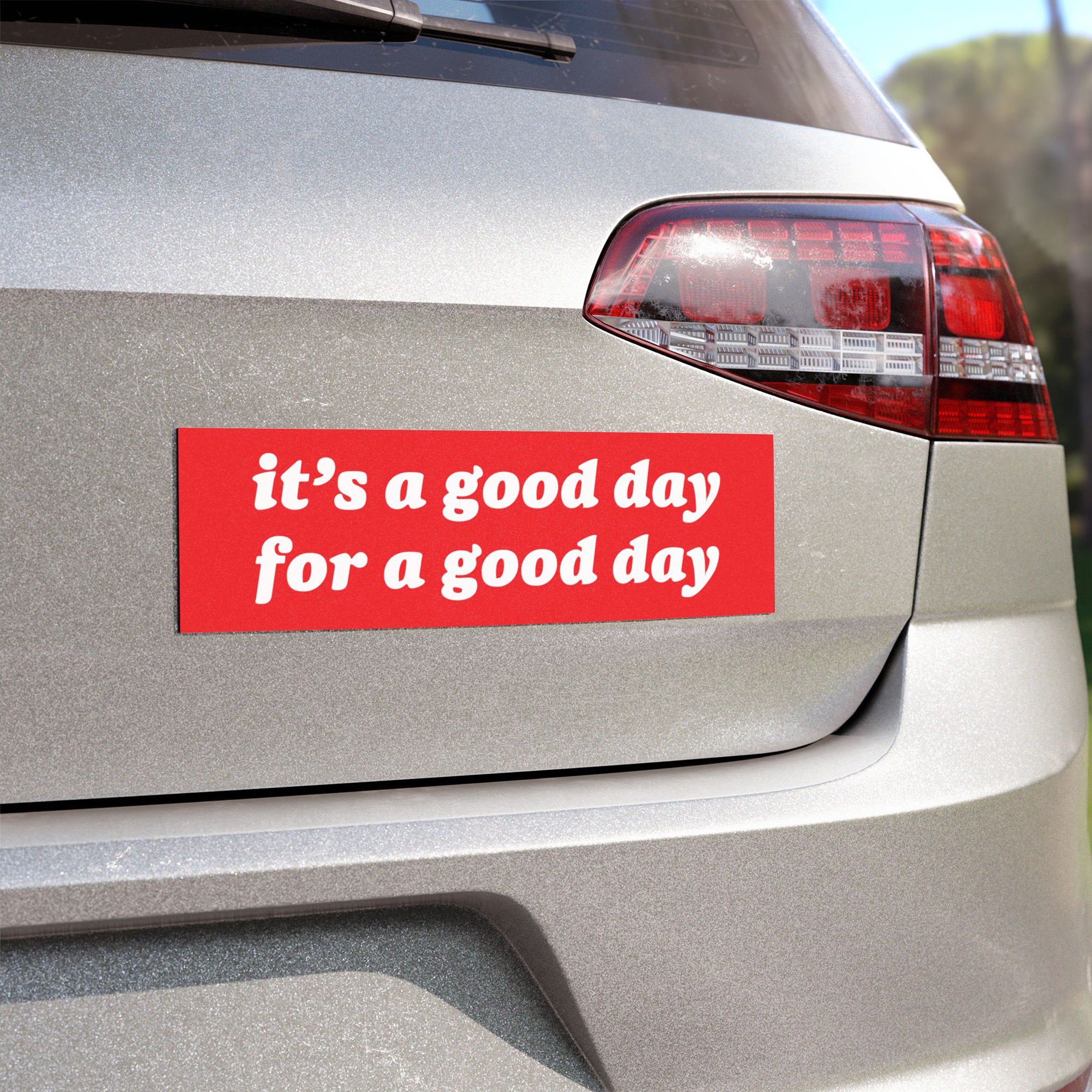 The rear of a silver car displays the Good Day Sticker & Magnet, featuring the motivational phrase it's a good day for a good day in white text on a red background.
