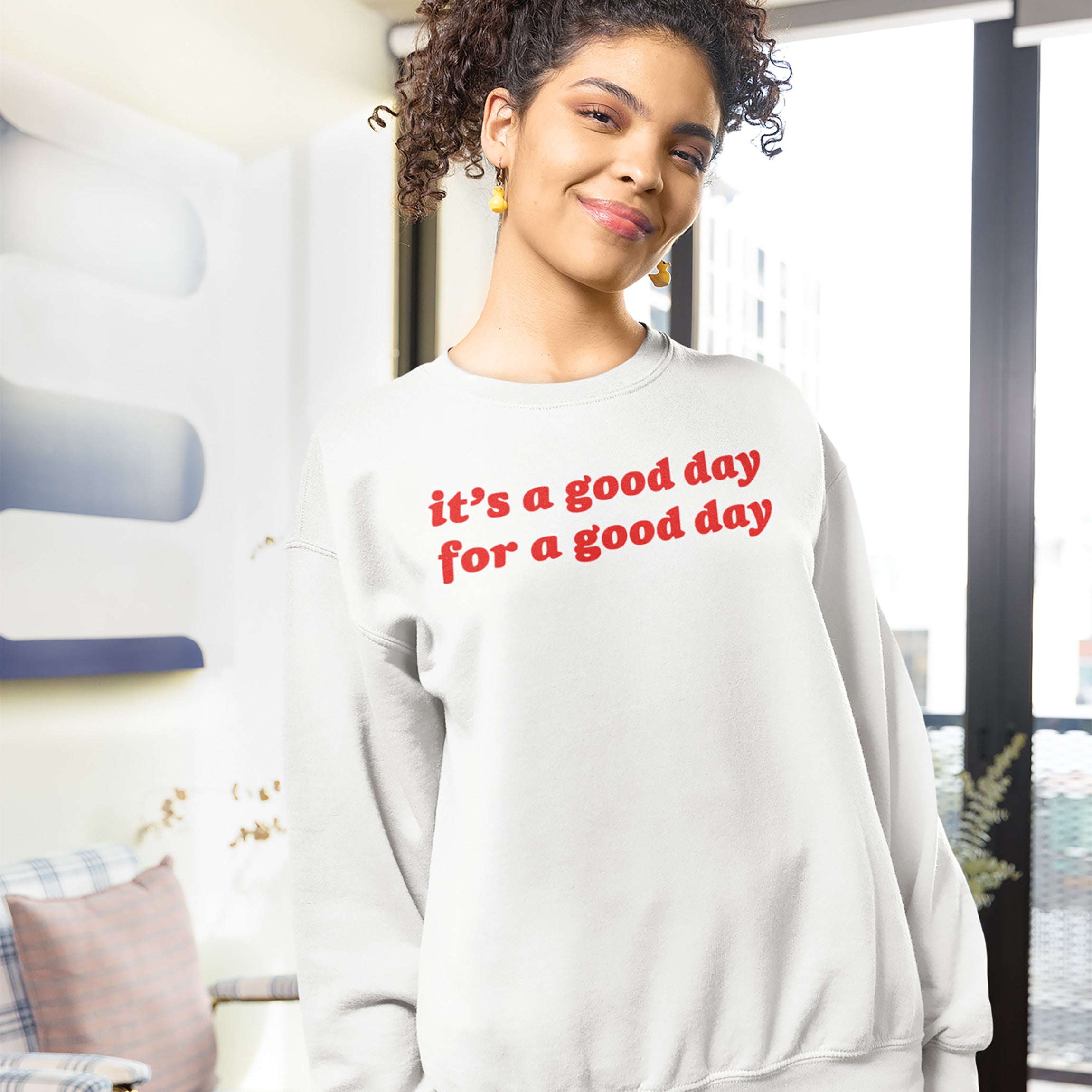 Young person indoors wearing the Good Day Shirt, a white sweatshirt with bold red text, “it’s a good day for a good day,” promoting positivity and mindfulness with vintage charm.