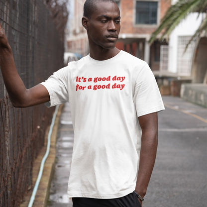 Young man leans on a fence wearing the Good Day T-shirt, featuring bold red script, “it’s a good day for a good day,” embodying optimistic vibes in a candid, urban setting.