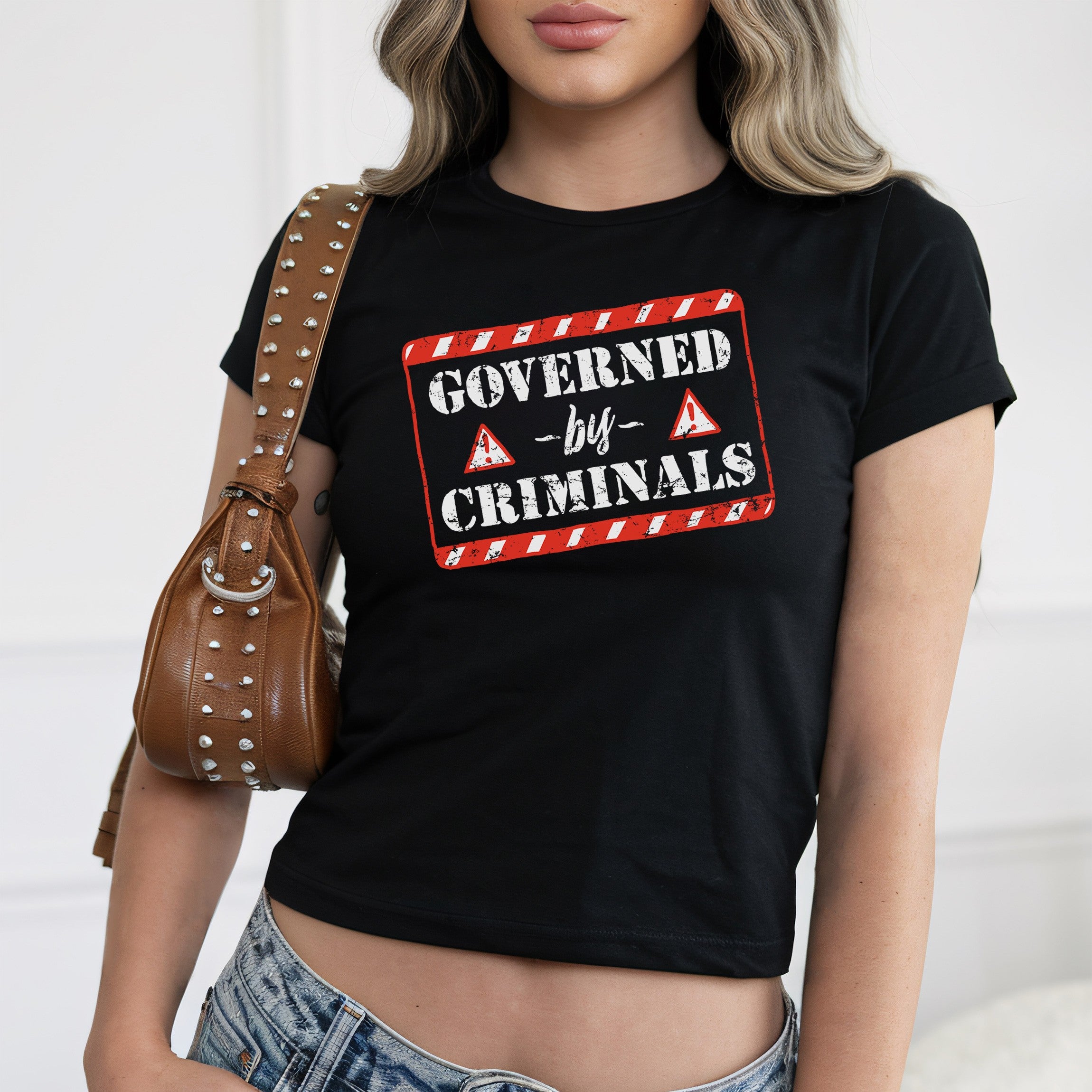 Governed By Criminals Women's Baby Tee features a bold, distressed sign design, perfect for making a rebellious statement. Pairing it with a studded shoulder bag and jeans adds a stylish edge.