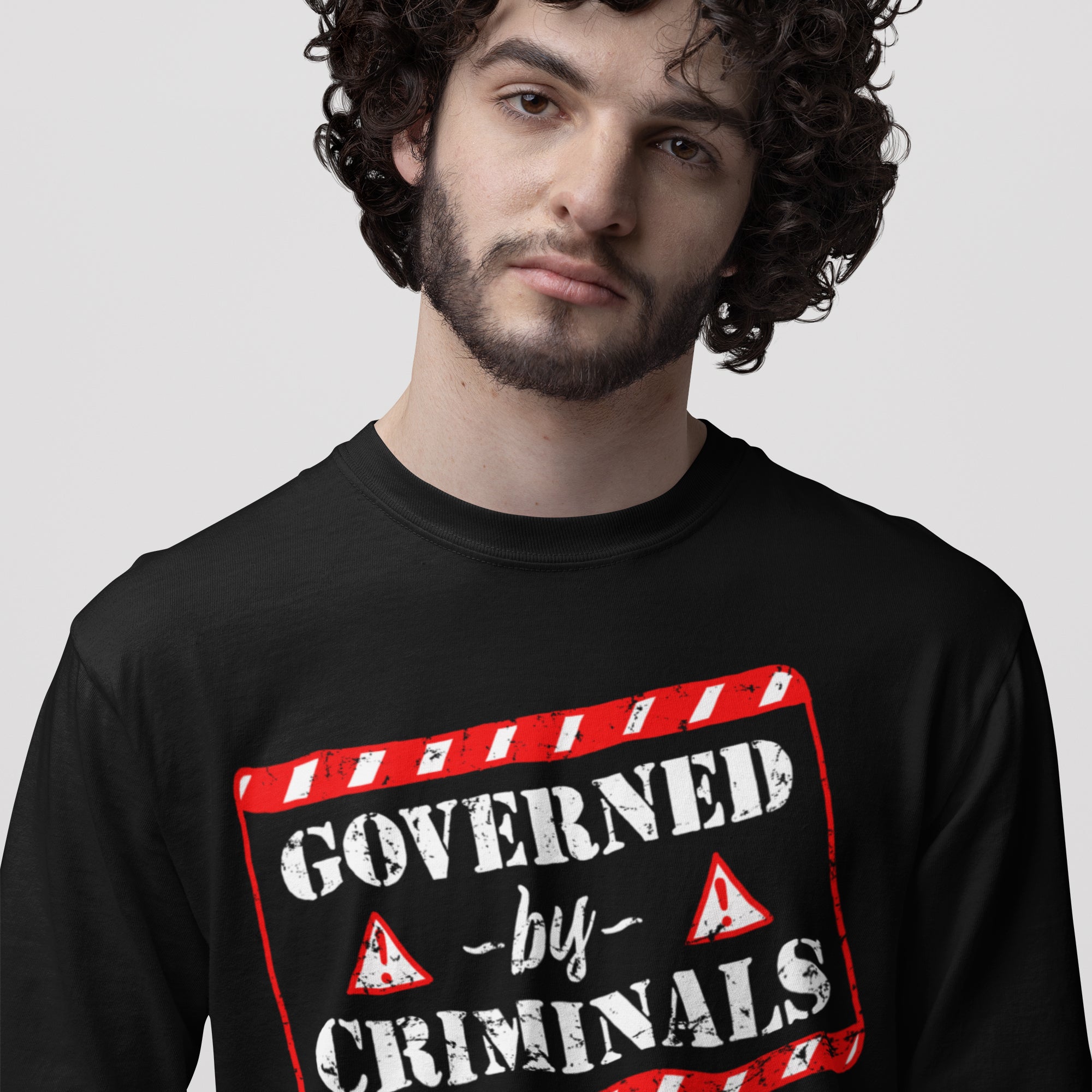 Young man in black long-sleeve Governed By Criminals Shirt featuring bold stencil-style text and red accents, symbolizing rebellion against political corruption and supporting social justice causes.