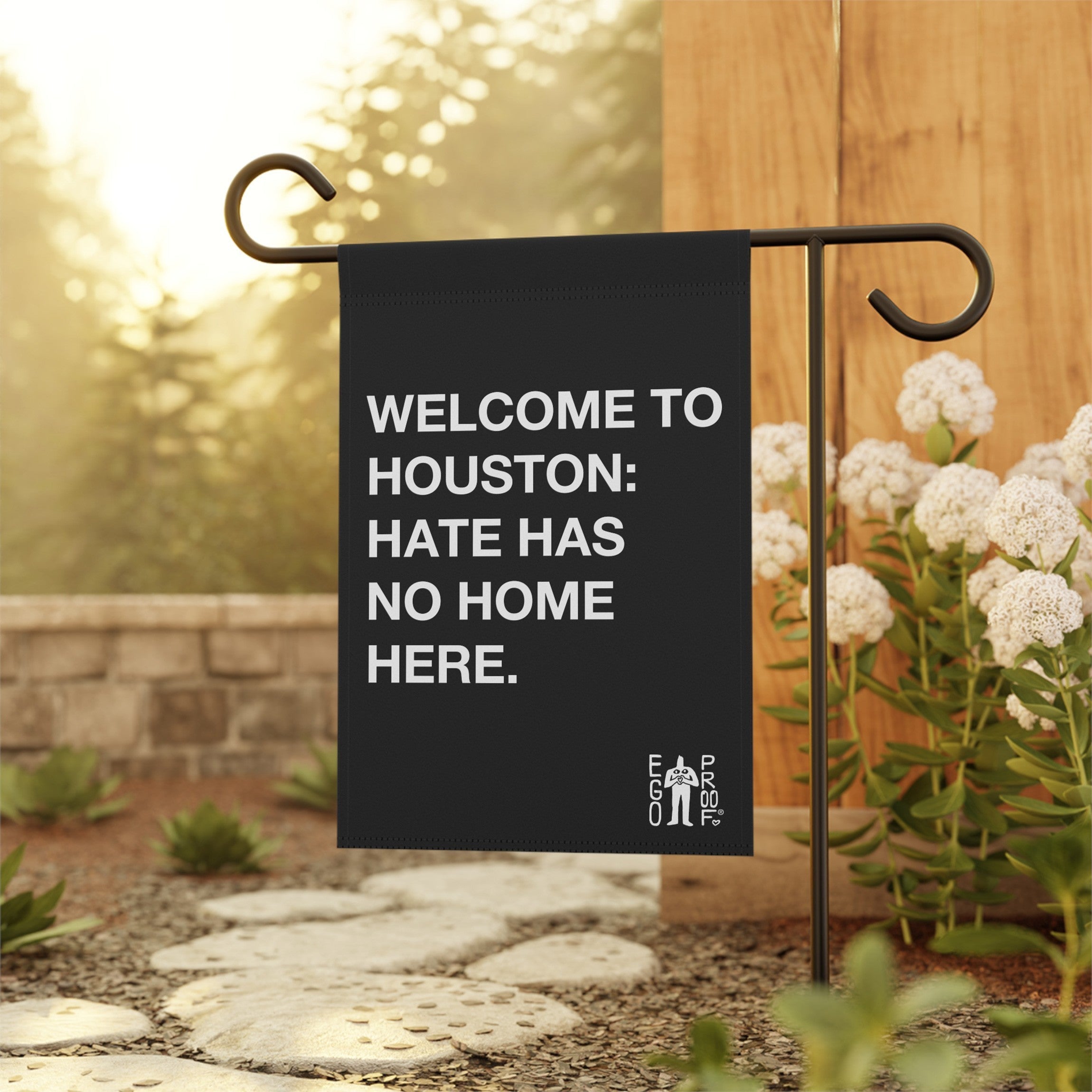 Custom Hate Has No Home Here Garden Flag displayed on a black metal stand by a stone pathway, surrounded by white flowers, promoting unity and inclusion with bold text against a natural backdrop.