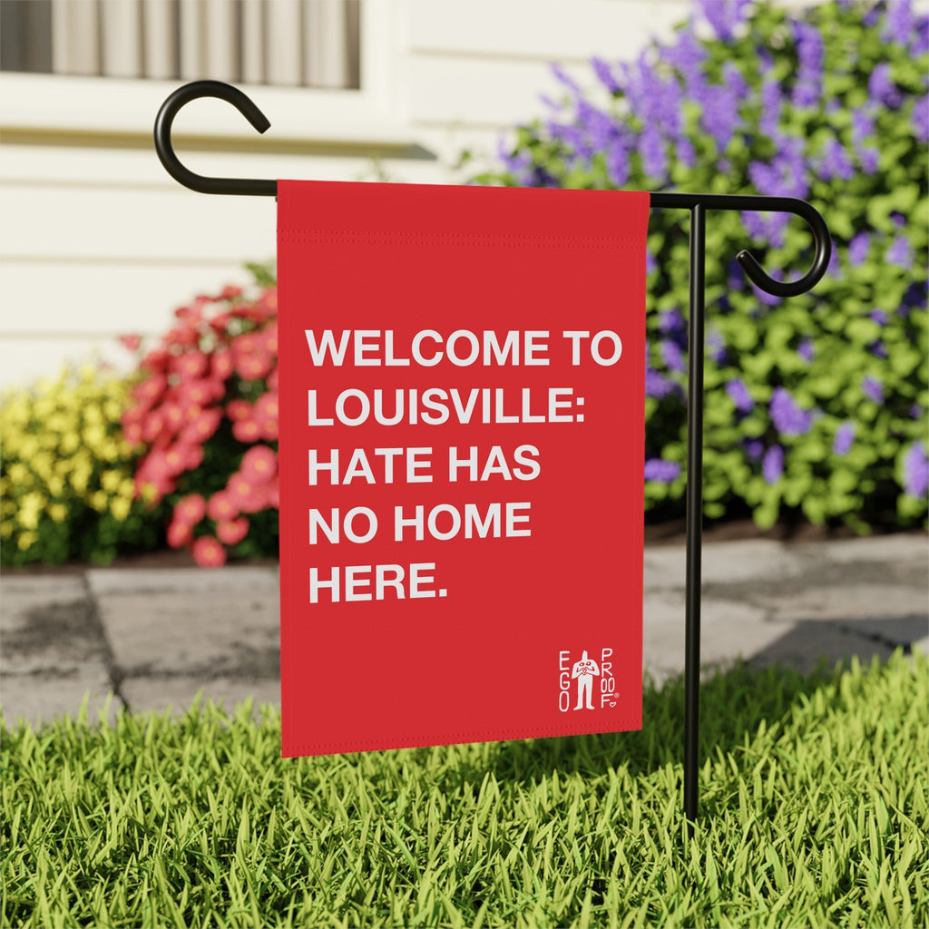 Custom Hate Has No Home Here Garden Flag staked in a lawn, displaying WELCOME TO LOUISVILLE: HATE HAS NO HOME HERE in white letters, surrounded by grass and flowering shrubs against a house backdrop.