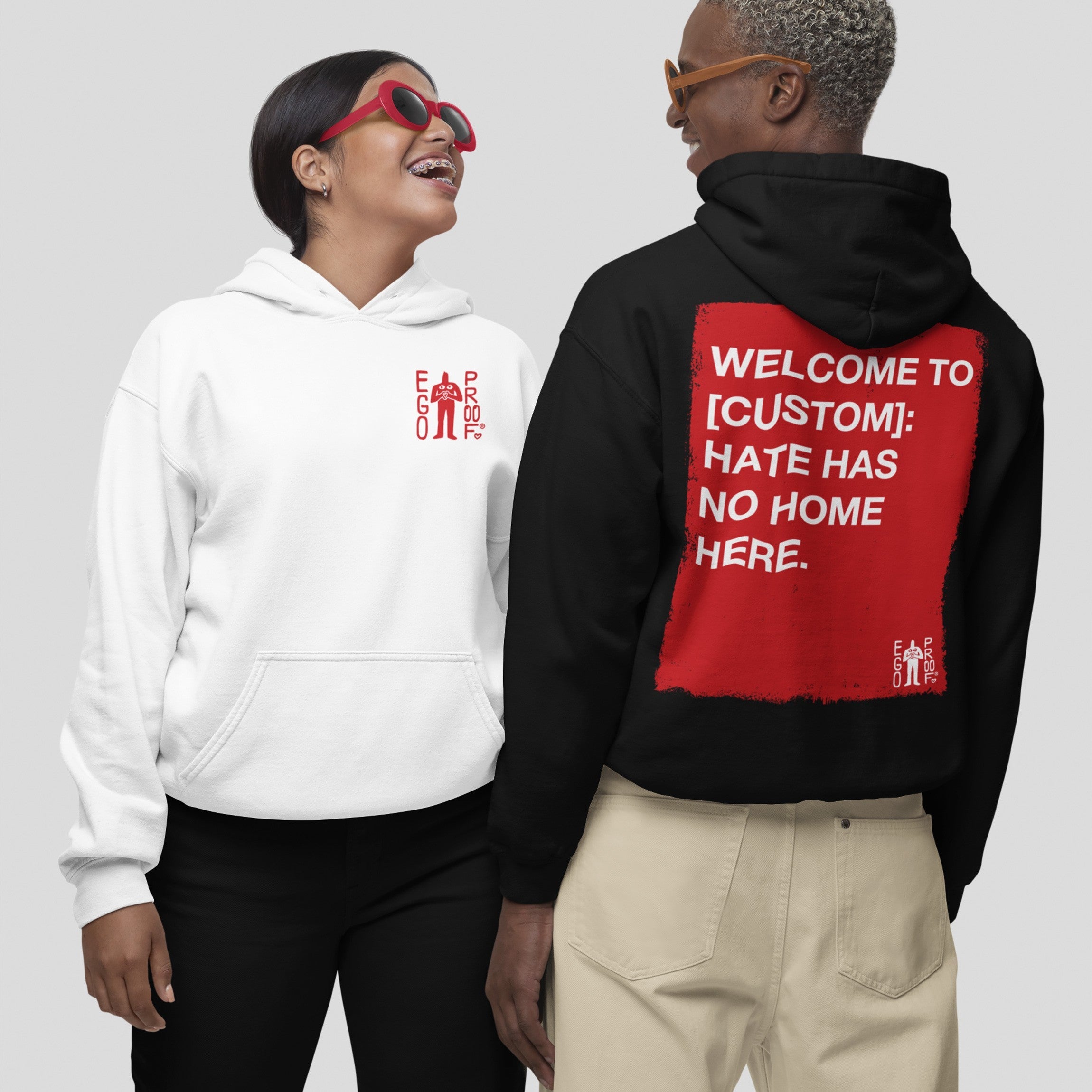 Two people wearing “Custom Hate Has No Home Here Hoodie”; one in a white hoodie with a small “EGO PROD.” logo, the other in a black hoodie featuring a bold, customizable community message on the back.