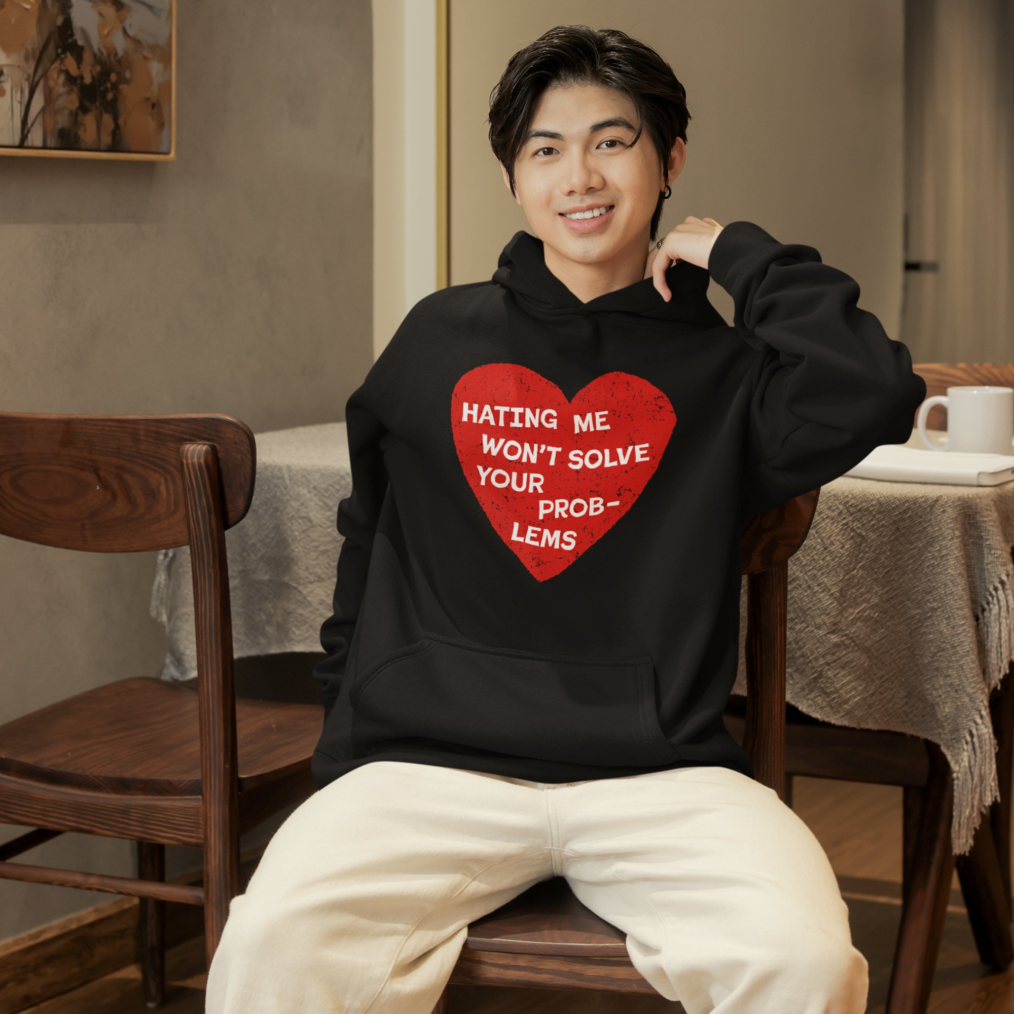 Young man in a cozy café wears the No Hate Shirt, a black hoodie with a bold red heart and the slogan “Hating me won’t solve your problems,” advocating love and equality.
