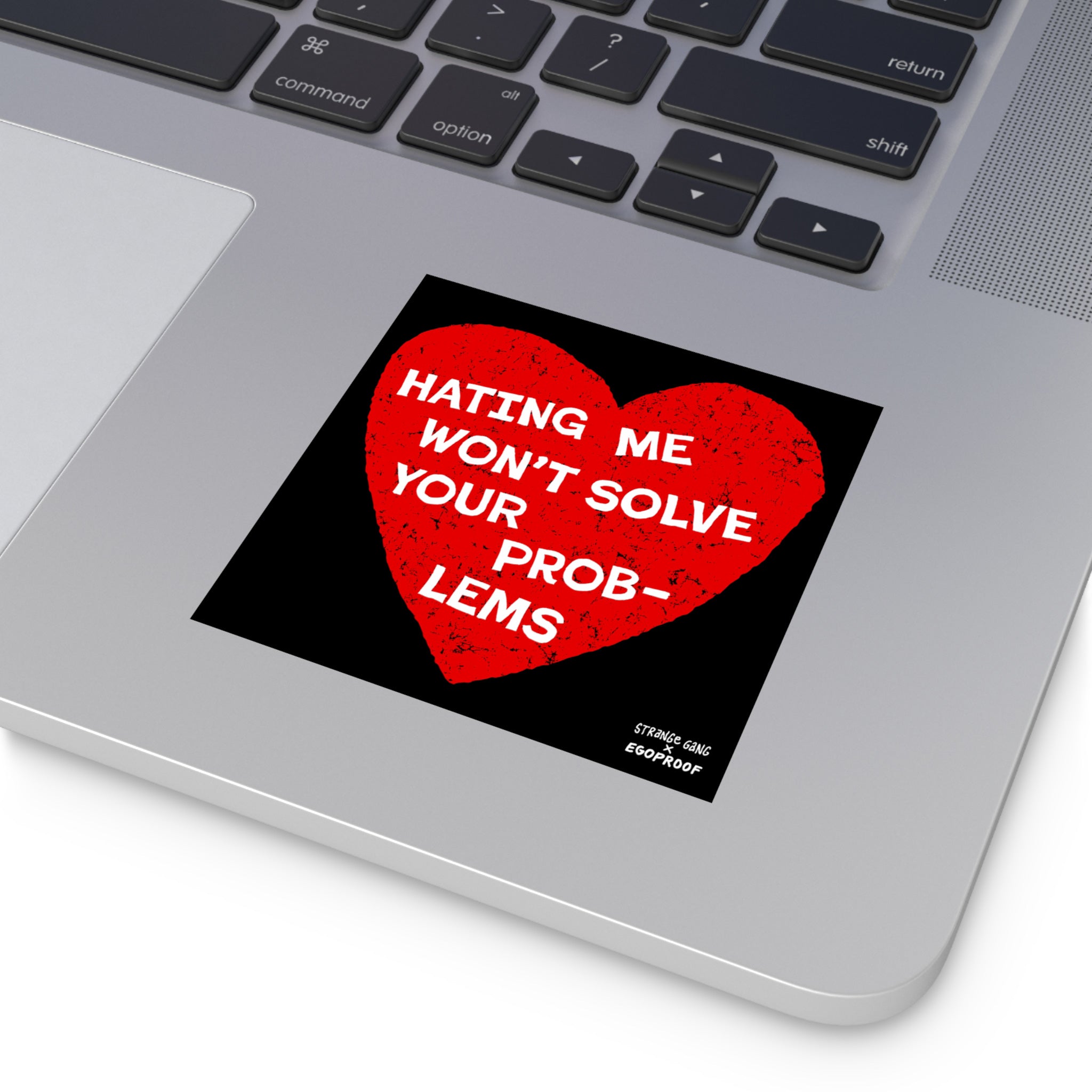 Silver laptop keyboard with a black square sticker below featuring a distressed red heart and white text: “Hating Me Won’t Solve Your Problems.” Includes credit: “STRNGE GANG × EGOPROOF.”