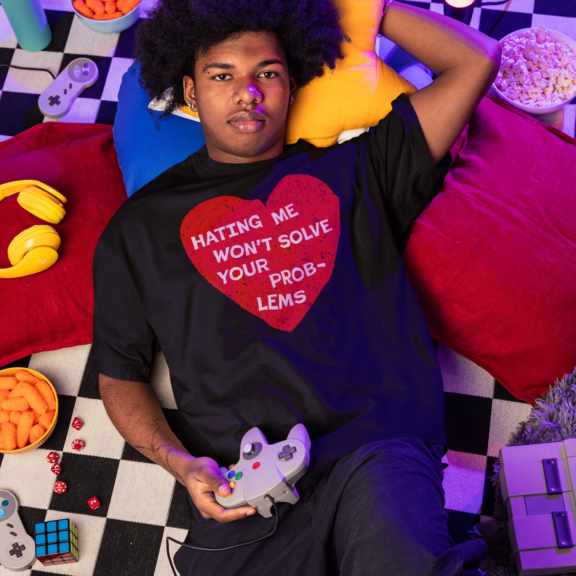 Young man on checkerboard blanket, holding a retro controller, wears the No Hate Shirt with a bold red heart message, surrounded by gaming gear and snacks in a cozy setting.