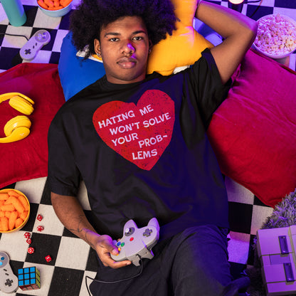 Young man on checkerboard blanket, holding a retro controller, wears the No Hate Shirt with a bold red heart message, surrounded by gaming gear and snacks in a cozy setting.