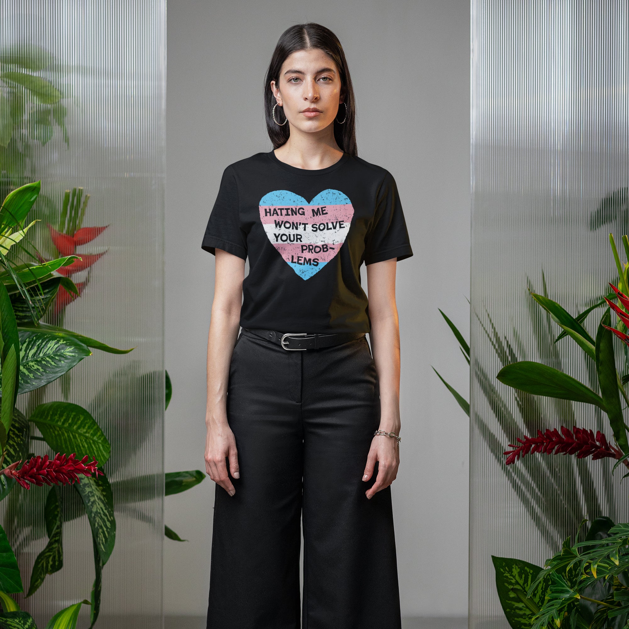 Person wearing the No Trans Hate Shirt featuring a trans pride heart with the words “Hating Me Won’t Solve Your Problems,” standing between translucent dividers with lush potted plants on either side.
