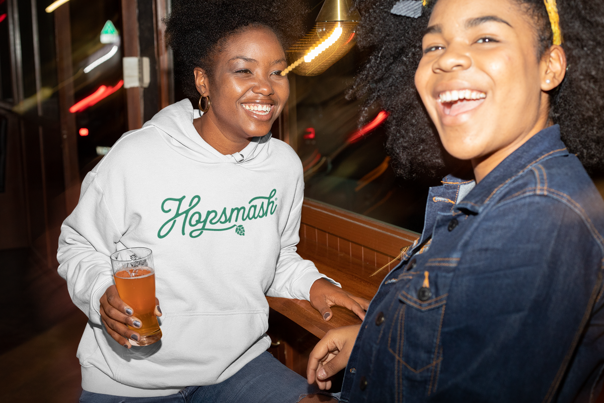 Two women smiling in a dimly lit bar or restaurant setting. The woman on the left wears a white hoodie with the word 'Hopsmash' in green script and holds a glass of amber beer.