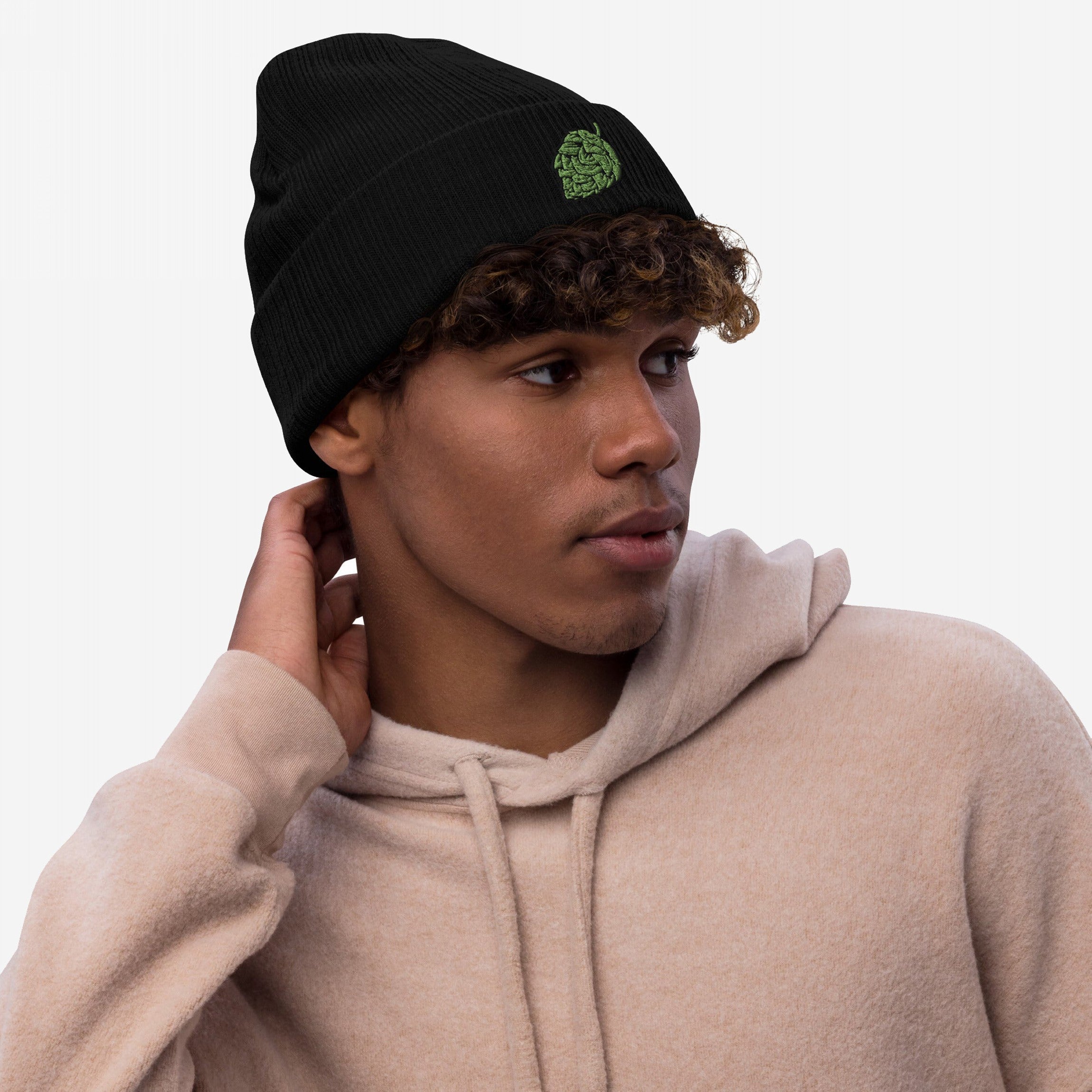 Young man in a beige hoodie models the Hop Nug Hat, a black rib-knit beanie with a green hop-cone motif, showcasing casual confidence for craft beer enthusiasts.