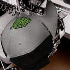 Hop Nug Hat in an embroidery machine, featuring a green hop-leaf design being stitched onto its gray front. Ideal for craft beer lovers who appreciate style and brewing culture.