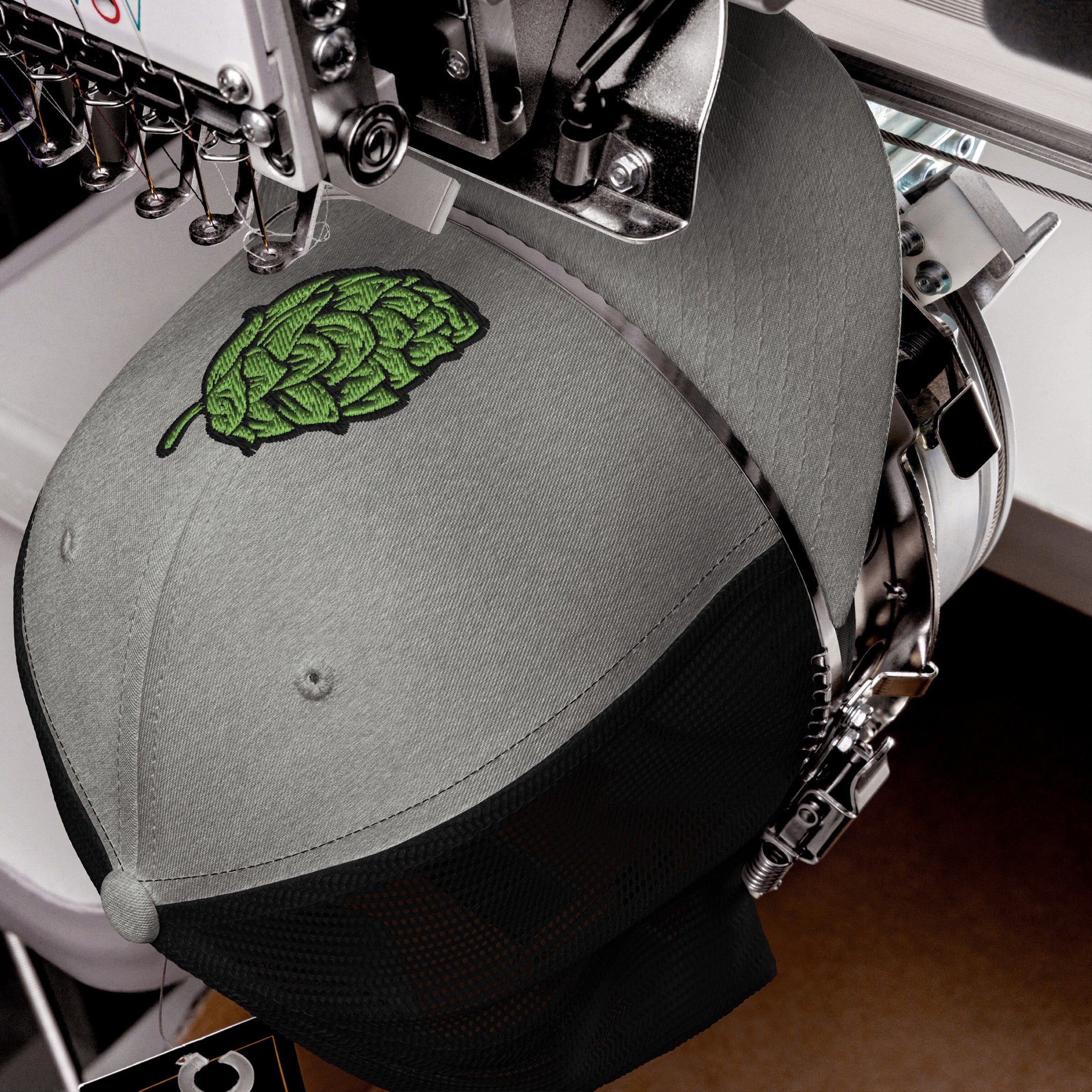 Hop Nug Hat in an embroidery machine, featuring a green hop-leaf design being stitched onto its gray front. Ideal for craft beer lovers who appreciate style and brewing culture.