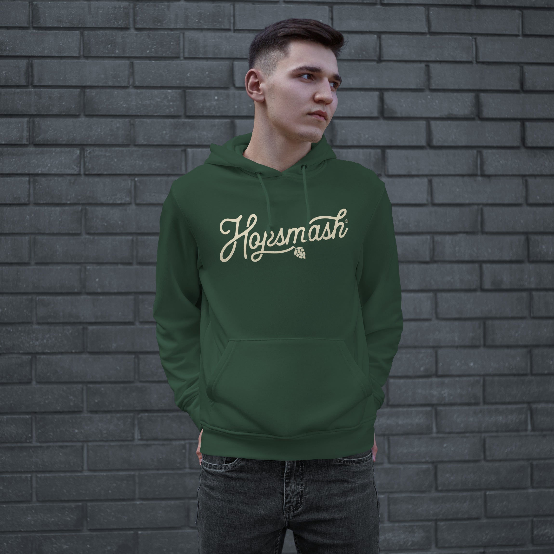 Young man wearing the Hopsmash Hoodie, featuring a forest-green pullover with Hopsmash script and hop-cone graphic, standing confidently, embodying a bold statement of hop appreciation.