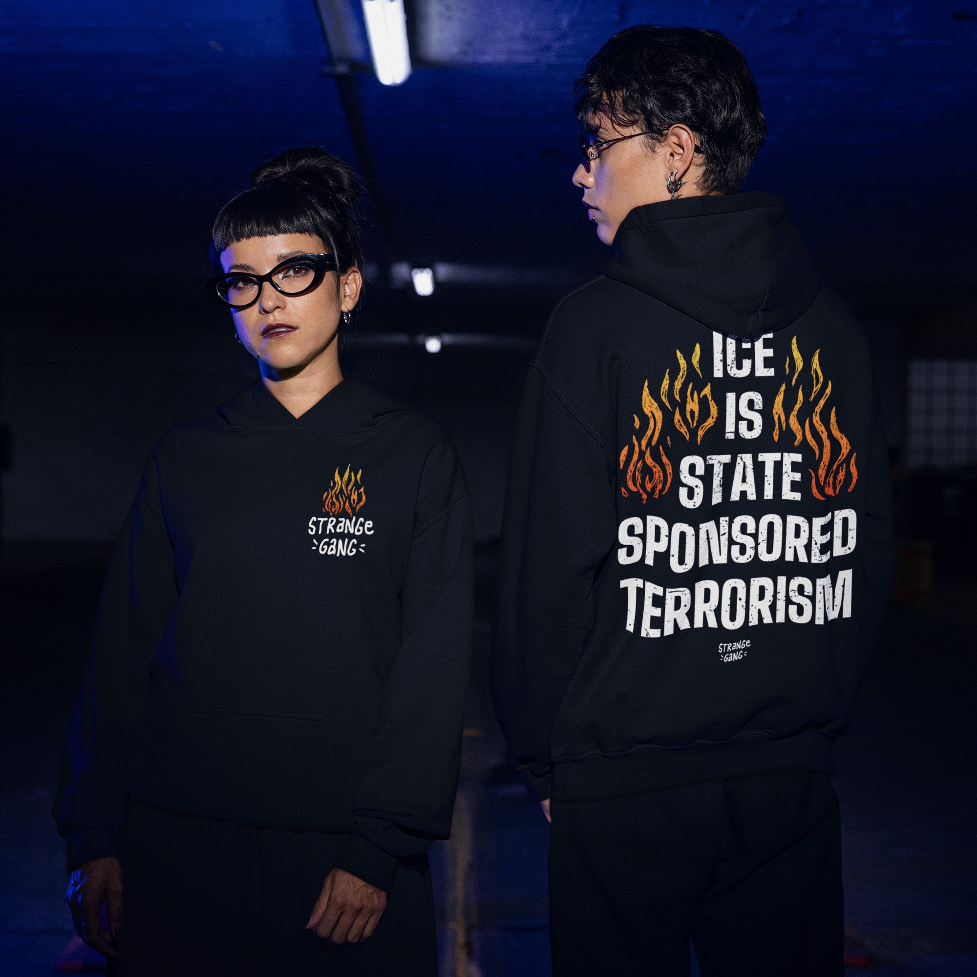 Two individuals in black hoodies; the left displays a small flame, while the right features ICE IS STATE SPONSORED TERRORISM in bold white text, highlighting the ICE Is State Sponsored Terrorism Shirt's activist message.