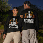 Two people stand outdoors at dusk wearing matching ICE Is State Sponsored Terrorism sweatshirts, each with a bold orange-flame graphic and white text, symbolizing a stance for immigrant rights.