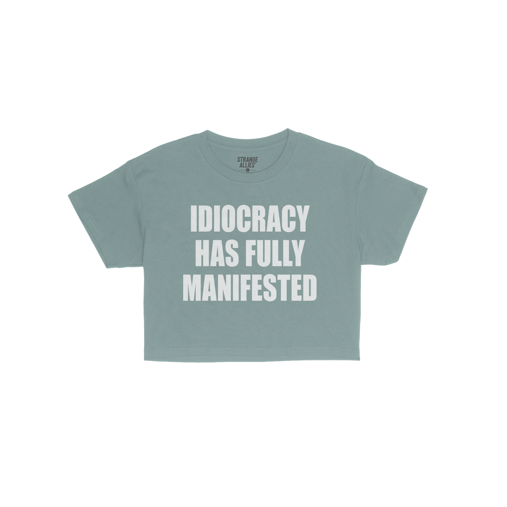 The Idiocracy Women's Baby Tee displays bold white text stating IDIOCRACY HAS FULLY MANIFESTED, capturing a satirical nod to 