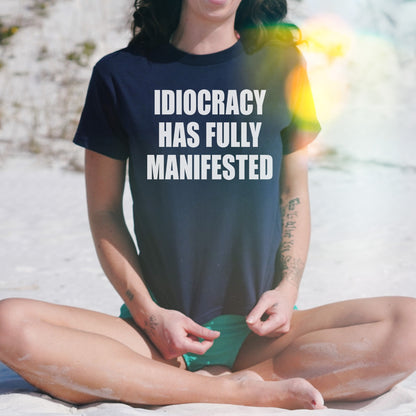 Person on beach wearing Idiocracy Women's Baby Tee, featuring IDIOCRACY HAS FULLY MANIFESTED text, paired with green swim bottoms, showcasing tattoos on forearms, with sunlight adding warmth to the scene.