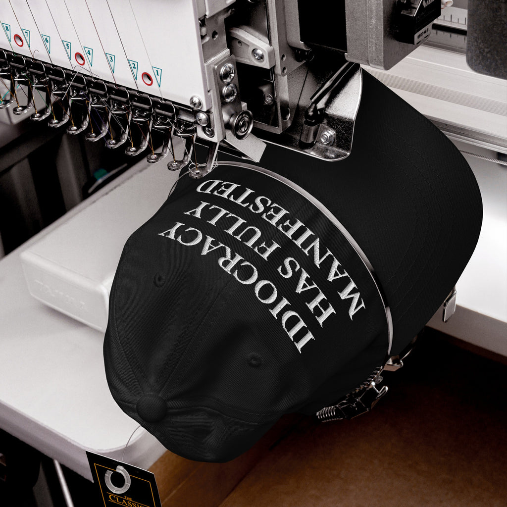 The Idiocracy Hat, captured mid-production, features a bold satirical statement being embroidered in white on a black cap, highlighting its classic serif typeface under a multi-needle embroidery machine.