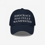 Navy-blue Idiocracy Hat with IDIOCRACY HAS FULLY MANIFESTED embroidered in white serif letters, reflecting bold social commentary and aligning with the store's advocacy for equality and justice.